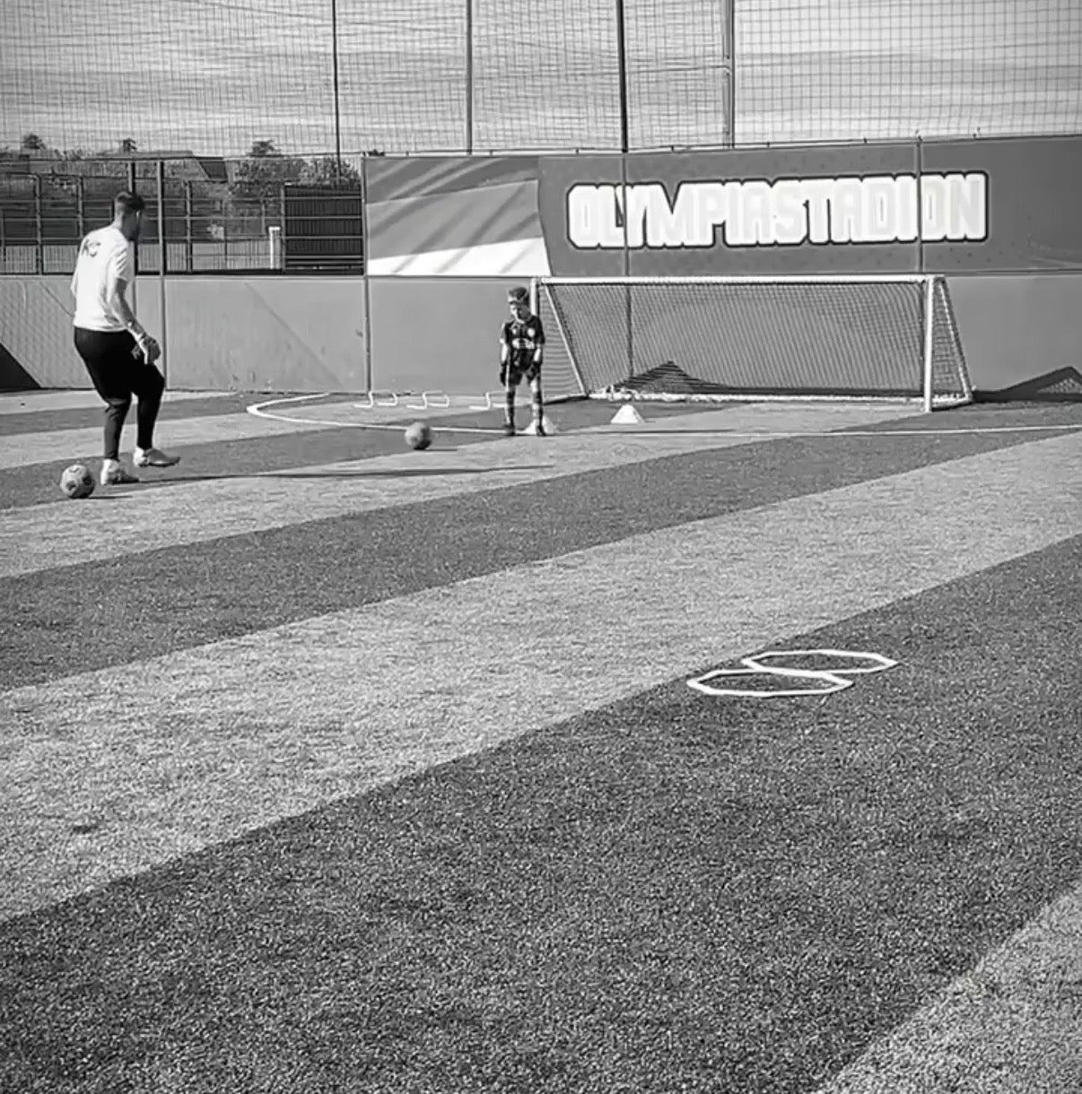 Black and white photo of a soccer training scene, showing a player kicking a ball on an artificial turf field with a goal in the background.