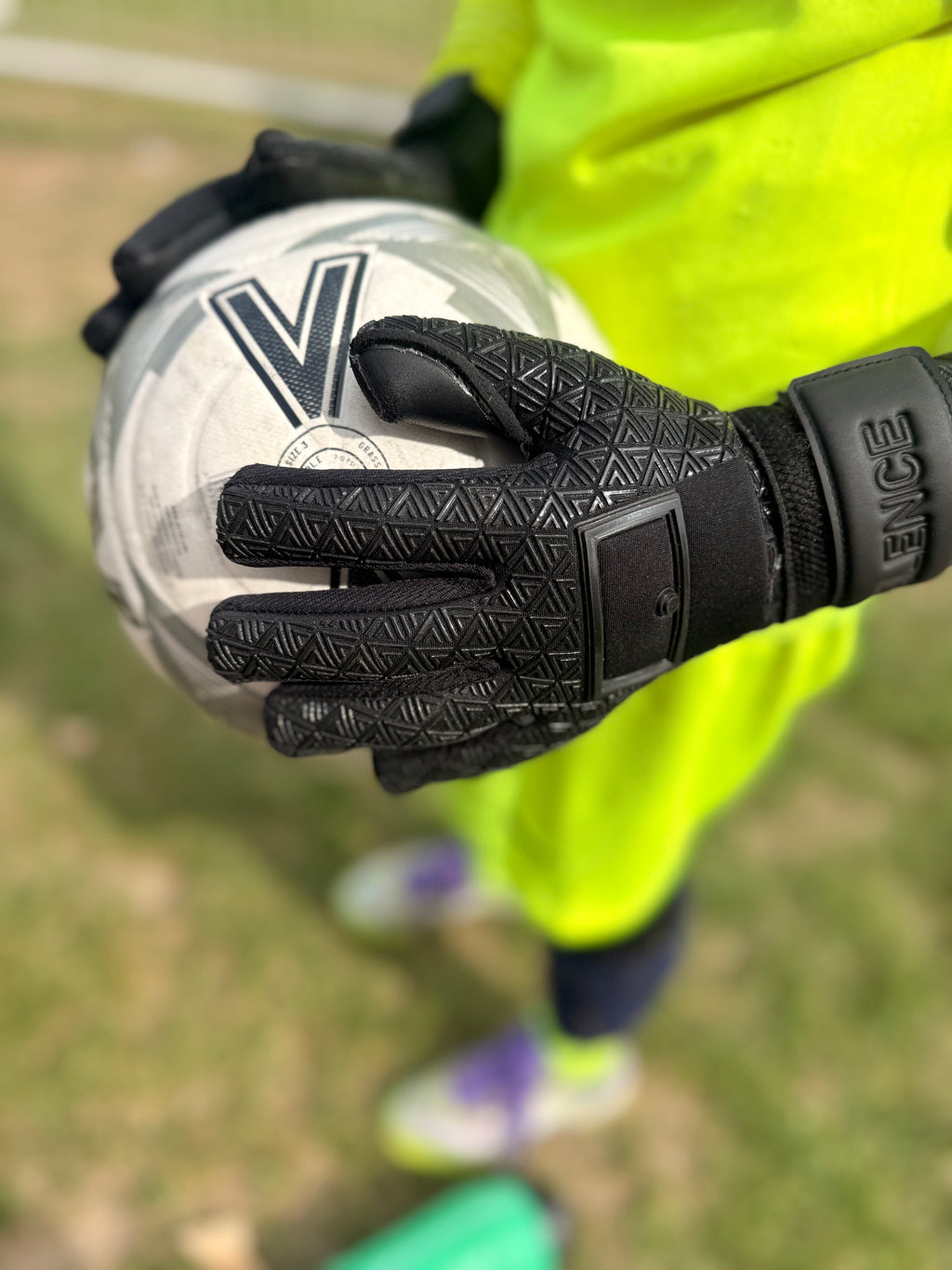 Foundation 01 Goalkeeper Gloves in black, holding a white soccer ball with a green field in the background