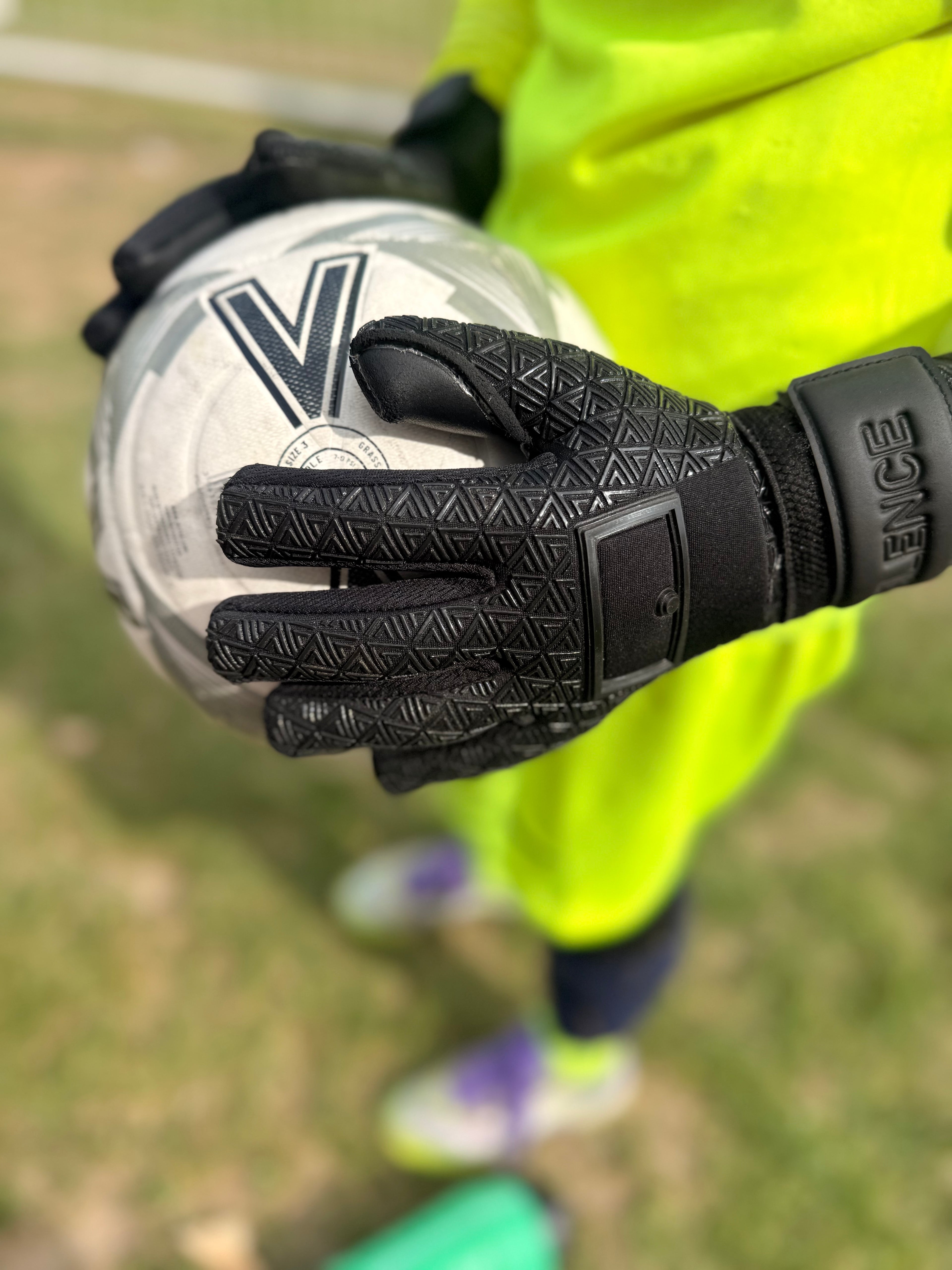 Foundation 01 Goalkeeper Gloves in black, holding a white soccer ball with a green field in the background